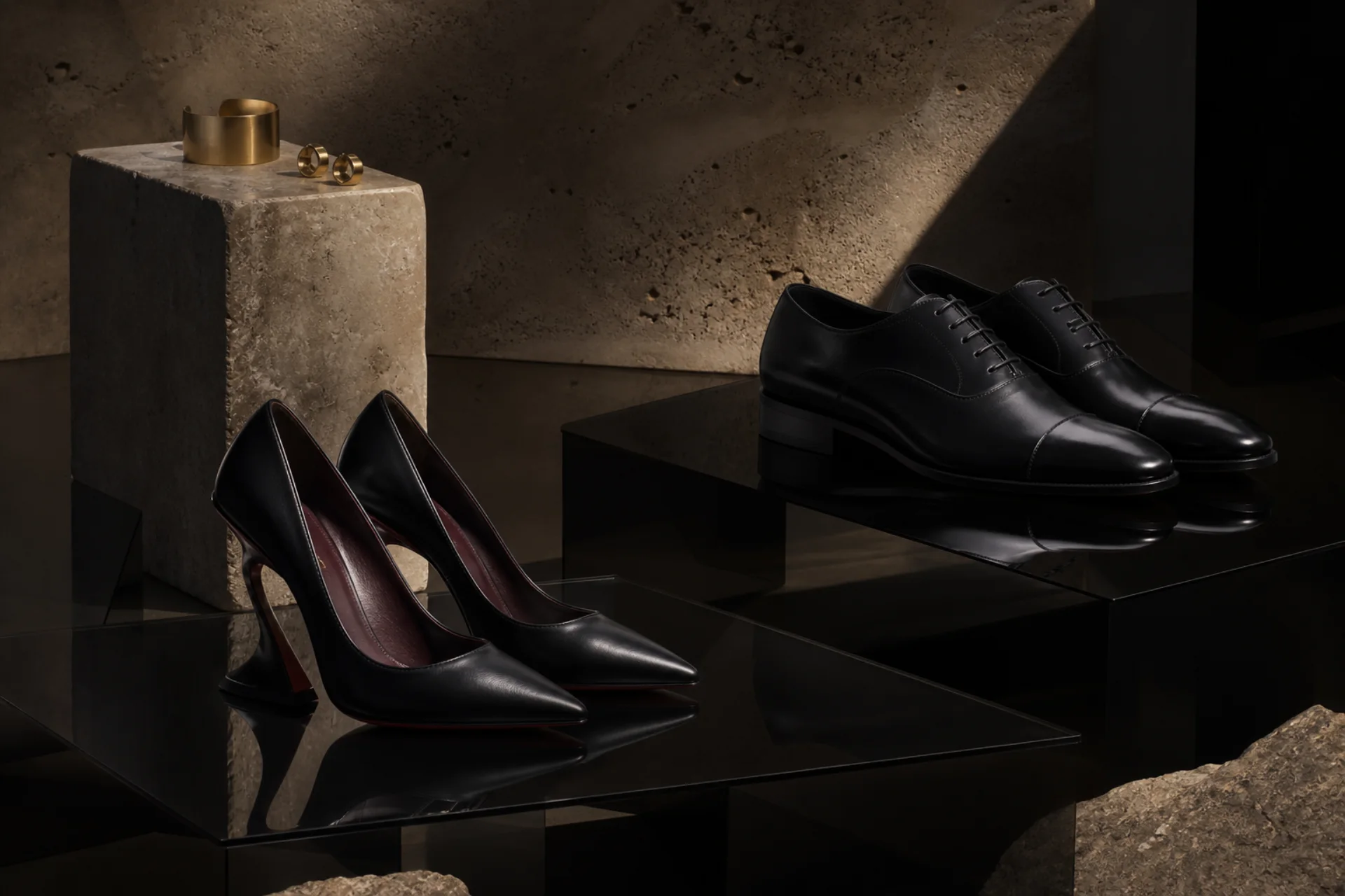Sculptural black footwear and gold jewelry displayed on stone and smoked glass